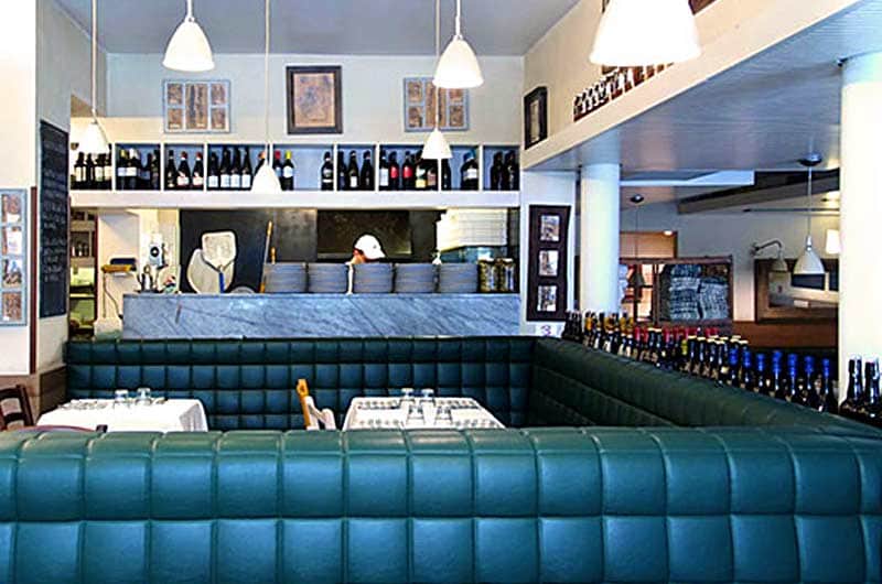 Sala con divanetti in pelle verde ad angolo, banco in marmo con piatti impilati e scaffali di bottiglie di vino alle pareti.