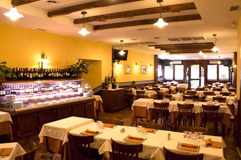Sala da pranzo con travi in legno a vista, tavoli con tovaglie bianche e lampade a sospensione.