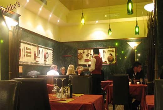Sala con pareti verdi, bottiglie verdi appese come lampade, un cameriere in gilet accanto a tavoli con tovaglie rosse.