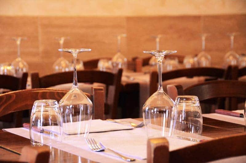 Calici da vino capovolti su tavoli apparecchiati con tovaglie bianche e sedie in legno scuro.