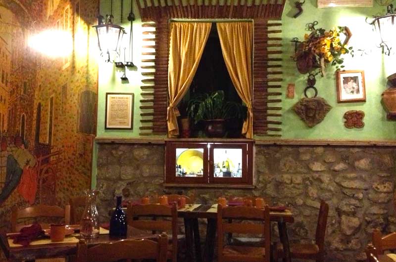 Sala con pareti verdi, tavoli in legno apparecchiati e una finestra con tende color senape su un muro in pietra.