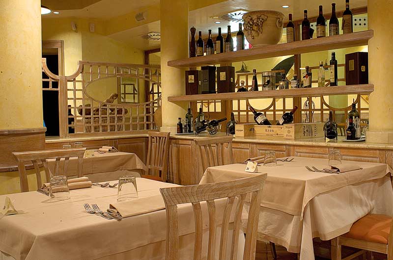 Sala con tavoli apparecchiati in tessuto beige e scaffali di bottiglie di vino sullo sfondo.