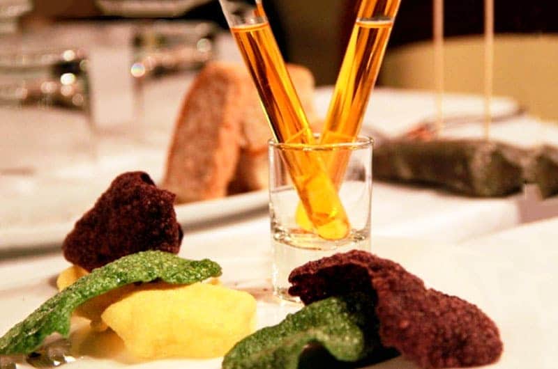 Due cannucce di vetro con liquido ambrato in un bicchierino, circondate da pezzi irregolari rossi, verdi e gialli su un piatto bianco.