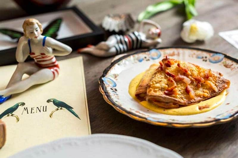 Crepe ripiegata con pancetta su crema gialla, accanto a un menu con due pappagalli disegnati e una statuina di donna in costume a righe.
