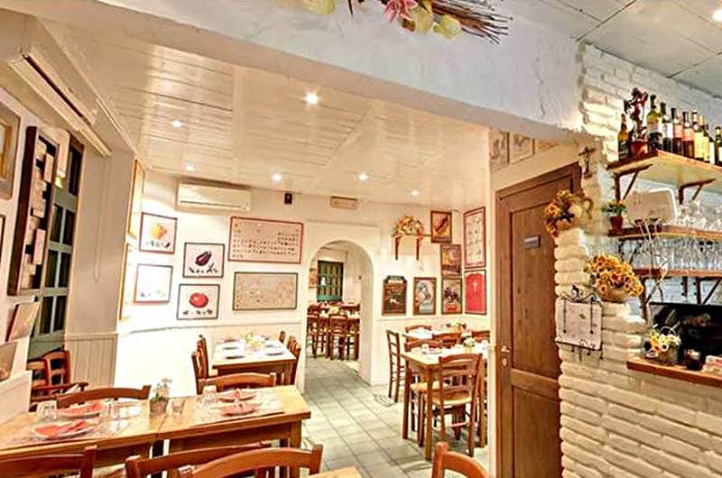 Sala da pranzo con tavoli in legno apparecchiati, pareti bianche con quadri e una parete in mattoni con mensole di bottiglie.