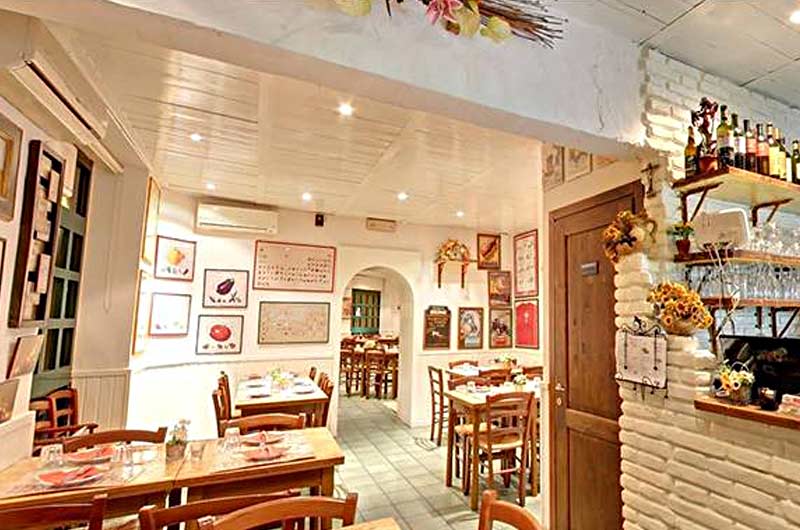 Sala da pranzo con tavoli in legno apparecchiati, pareti bianche con quadri e una parete in mattoni con mensole di bottiglie.