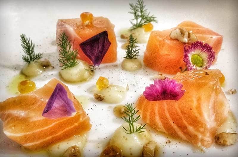 Cubi e tranci di salmone crudo su piatto bianco, con fiori edibili viola e rosa, rametti di aneto e piccole quenelle di crema.
