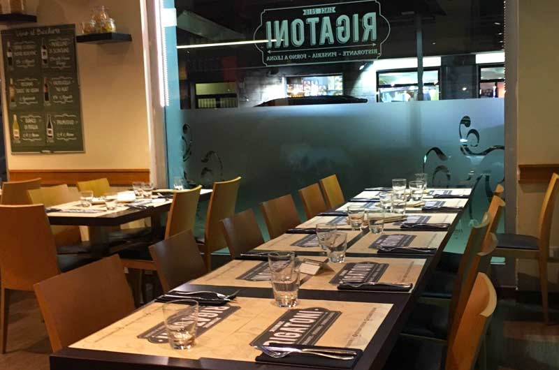 Fila di tavoli apparecchiati con bicchieri e set da tavola, davanti a una vetrata con insegna Rigatoni.