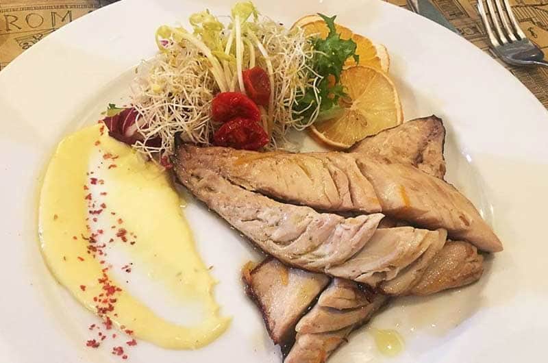 Filetti di pesce grigliato con germogli, pomodorini secchi e una salsa gialla decorata con polvere rossa.