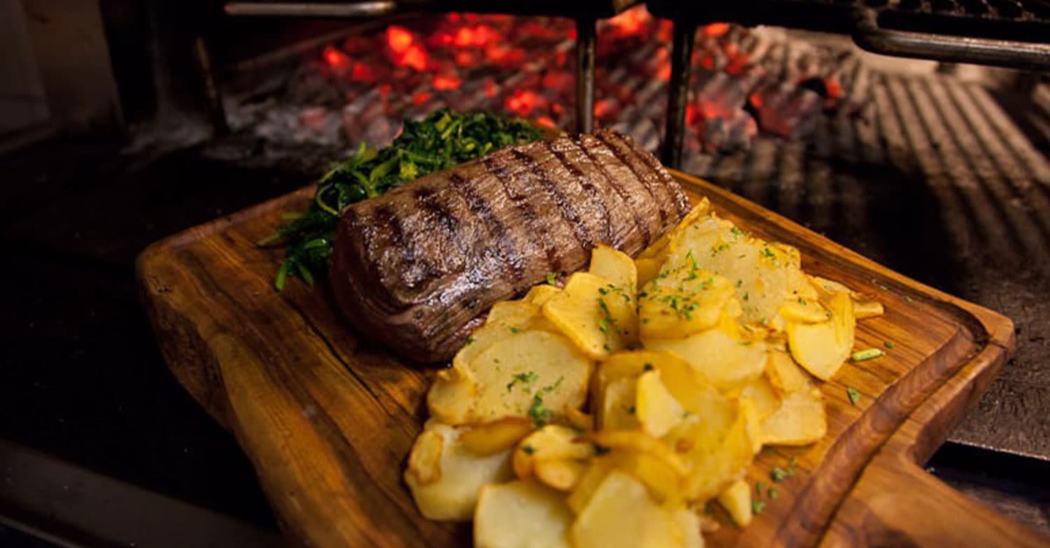 Carne grigliata a fette su tagliere di legno, con patate a spicchi e verdure a foglia verde, davanti a una griglia a carbone.