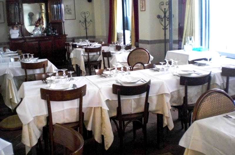 Sala di ristorante con tavoli apparecchiati di bianco e sedie in legno dallo schienale in paglia intrecciata.
