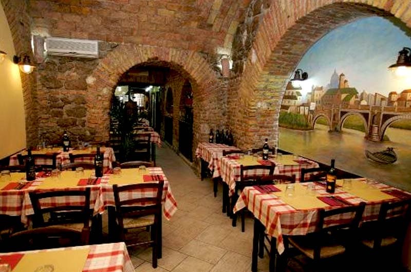 Sala con soffitto a volta in mattoni, tavoli con tovaglie a quadri rossi e un murale di paesaggio urbano sulla parete di destra.