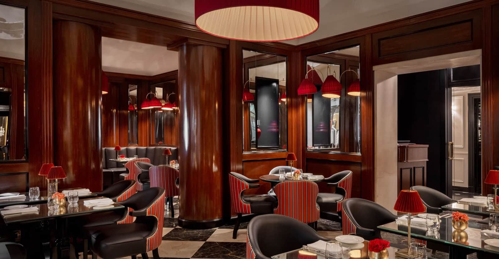 Sala con colonne in legno lucido, poltroncine a righe rosse e specchi alle pareti, illuminata da lampade a paralume rosso.