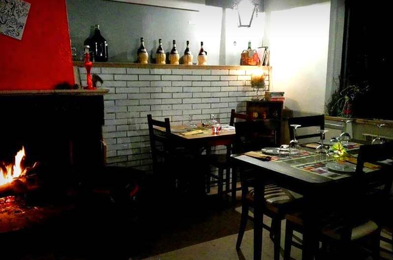 Sala da ristorante con camino acceso, muro in mattoni bianchi e tavoli apparecchiati con bicchieri da vino.