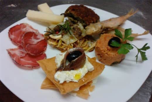 Piatto bianco con affettato rosato, cubi di formaggio, una frittura con erbe, un gambero fritto e una cialda con crema e acciuga.