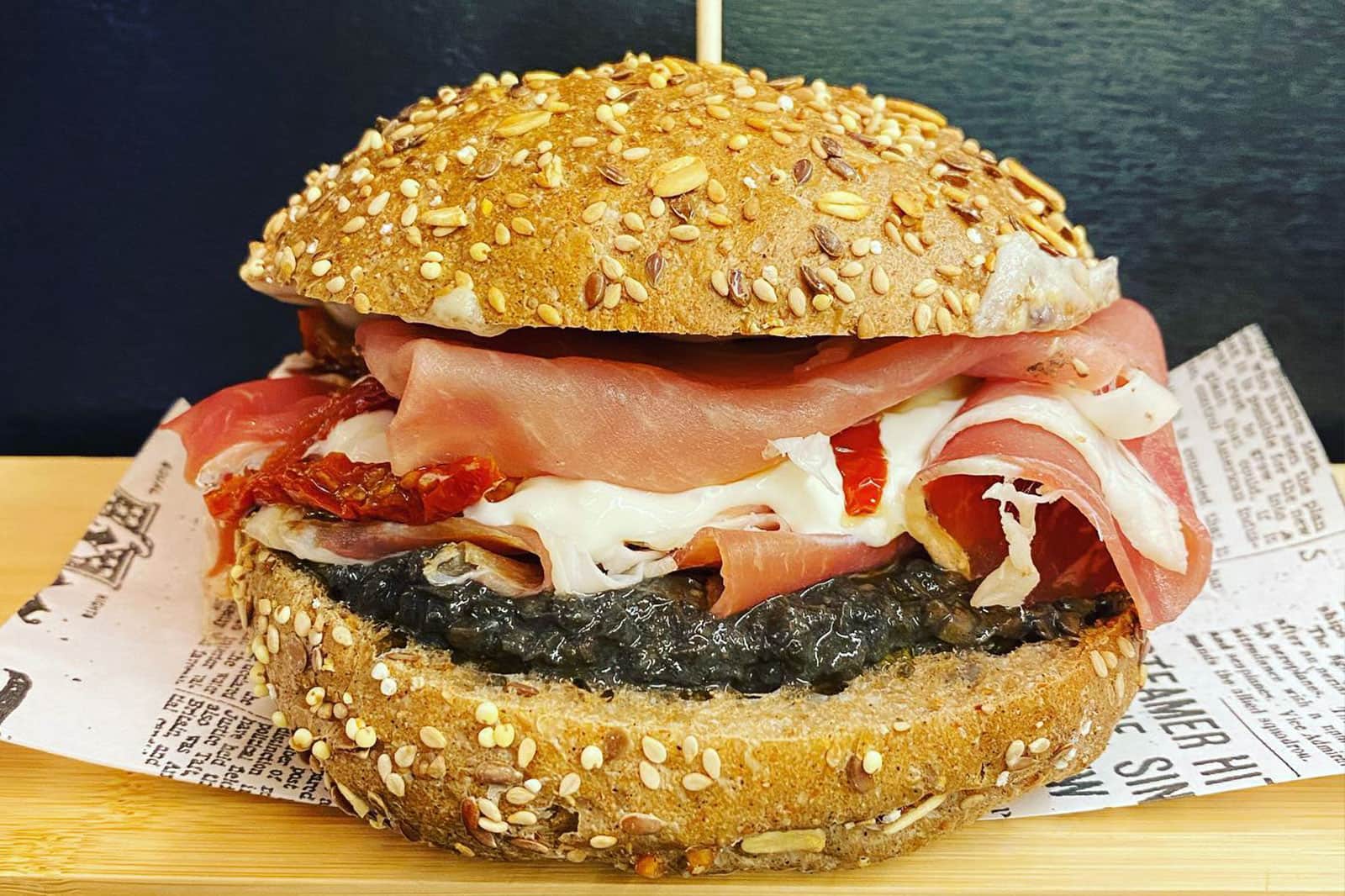 Panino con pane ai semi farcito di crema scura, mozzarella filante, pomodori secchi e fette di prosciutto crudo.