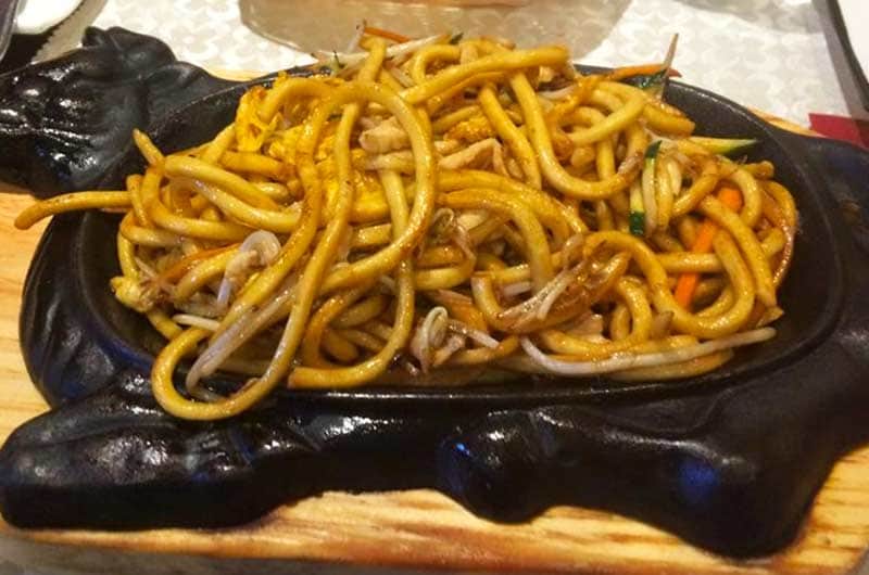 Noodles spessi conditi con salsa scura, germogli di soia e carote a julienne, serviti su piastra di ghisa nera.