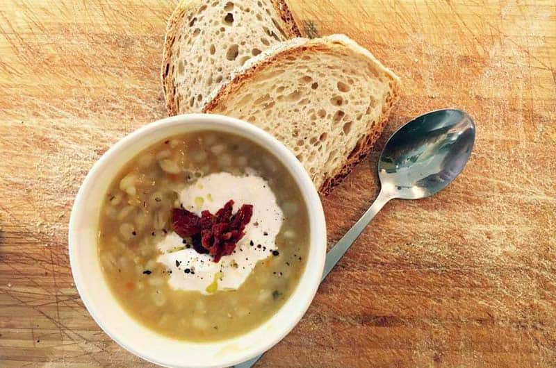 Ciotola di zuppa di legumi con crema bianca e pomodori secchi, accanto a fette di pane e un cucchiaio su un tagliere di legno.
