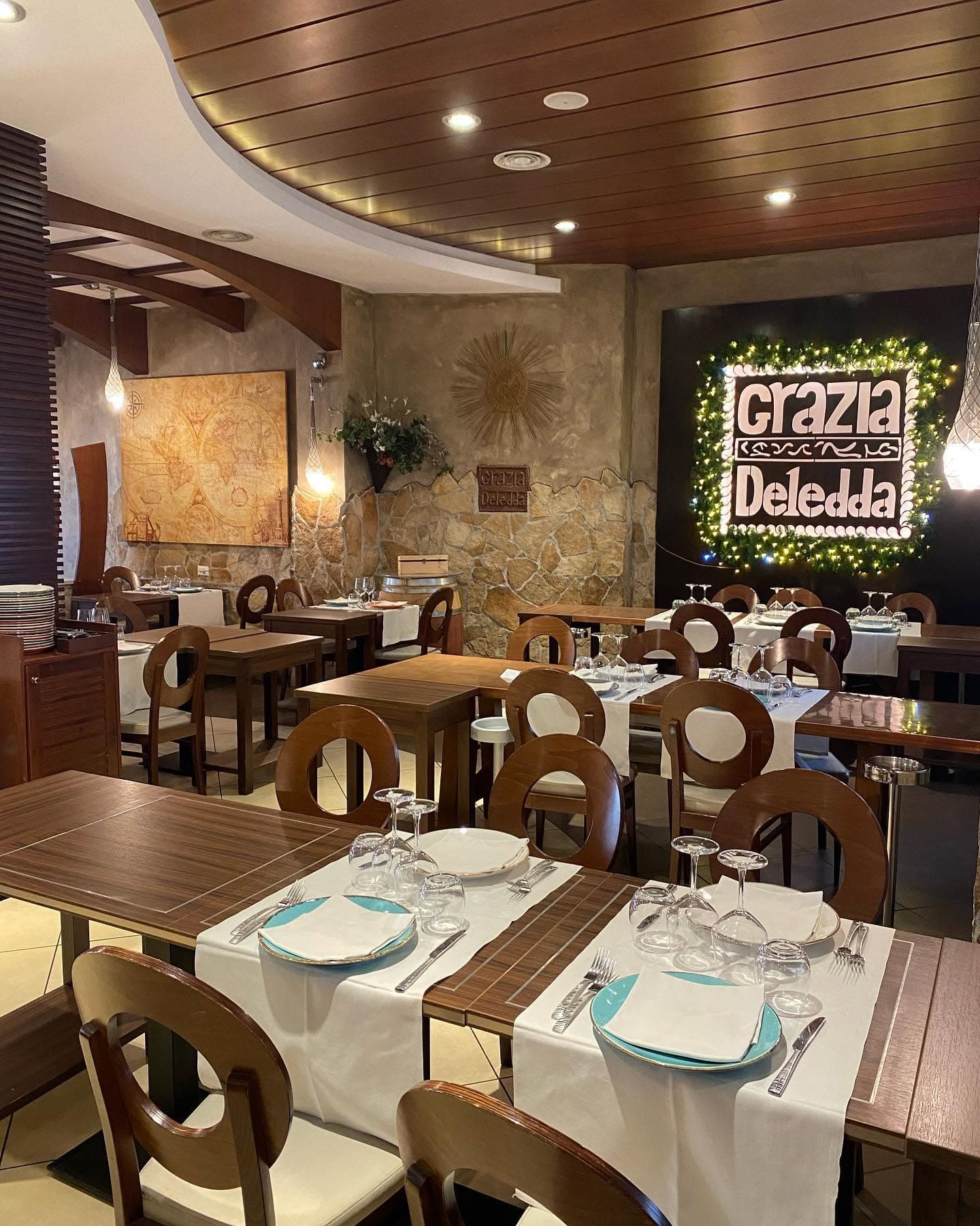 Sala con tavoli in legno apparecchiati e insegna luminosa 'Grazia Deledda' incorniciata da rami verdi.