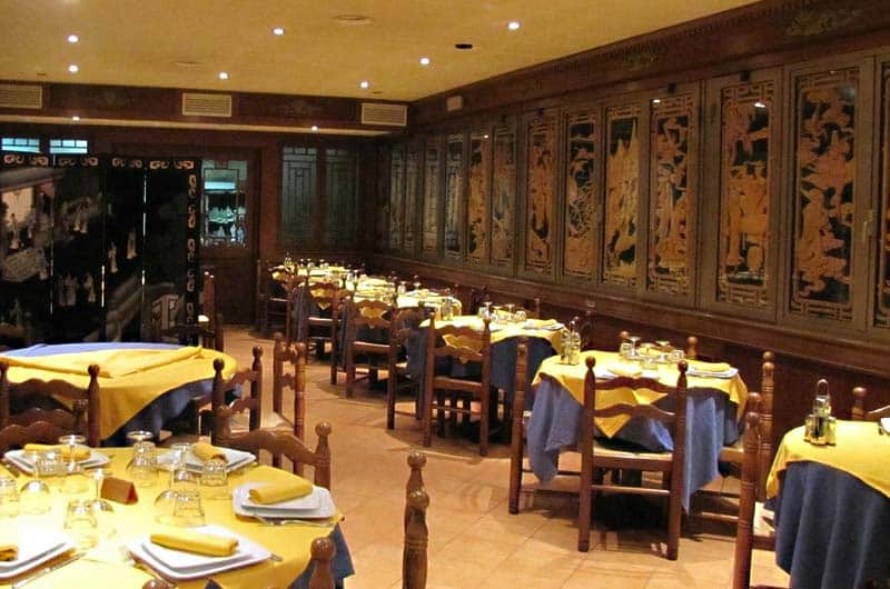 Sala da pranzo con tavoli apparecchiati di giallo e blu, pareti rivestite da pannelli intagliati dorati su fondo scuro.