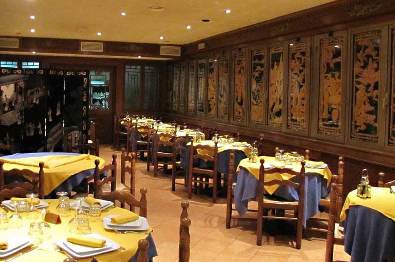 Sala da pranzo con tavoli apparecchiati di giallo e blu, pareti rivestite da pannelli intagliati dorati su fondo scuro.