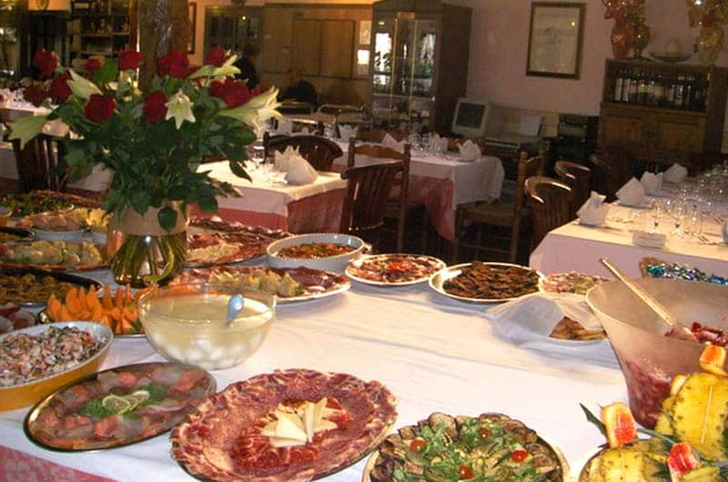 Tavolo buffet con vassoi di affettati e antipasti, vaso di rose rosse e gigli bianchi, sala con tavoli apparecchiati sullo sfondo.