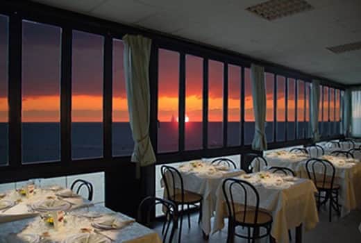 Sala con tavoli apparecchiati vicino a grandi vetrate, tramonto arancione sul mare sullo sfondo.
