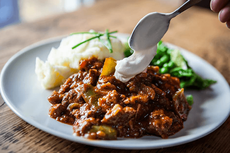 Cucchiaio versa panna acida su uno spezzatino di carne al sugo, con purè e fagiolini nel piatto.