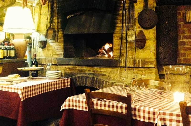 Sala con forno a legna acceso in un camino di mattoni, tavoli con tovaglie a quadri rossi e bianchi.