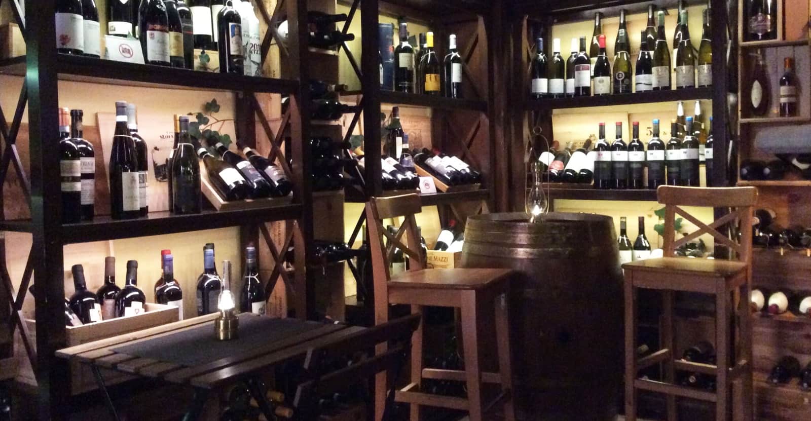 Scaffali in legno pieni di bottiglie di vino illuminate, con un barile e sedie al centro della sala.