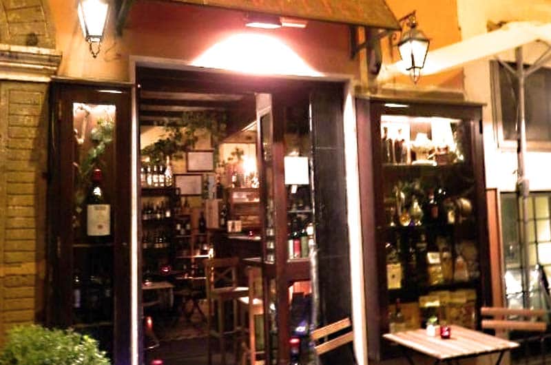 Ingresso illuminato di sera, con scaffali di bottiglie visibili all'interno e lampioni ai lati della porta.