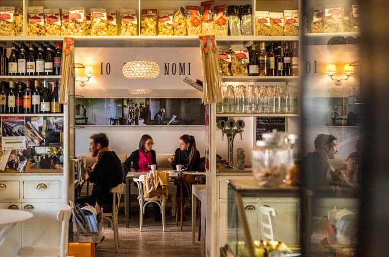 Sala di un ristorante con scaffali di vini e paste sopra un arco, tavoli con clienti seduti al centro.