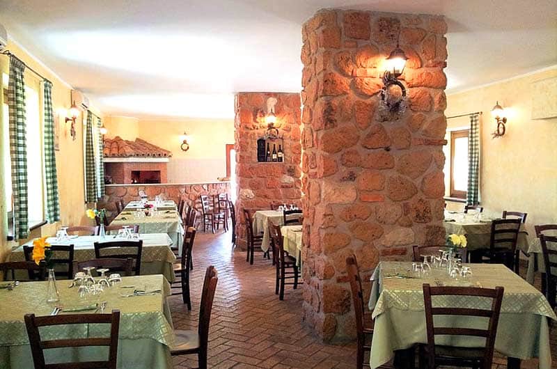 Sala da ristorante con pilastro in pietra al centro e tavoli apparecchiati con tovaglie chiare tra sedie in legno scuro.