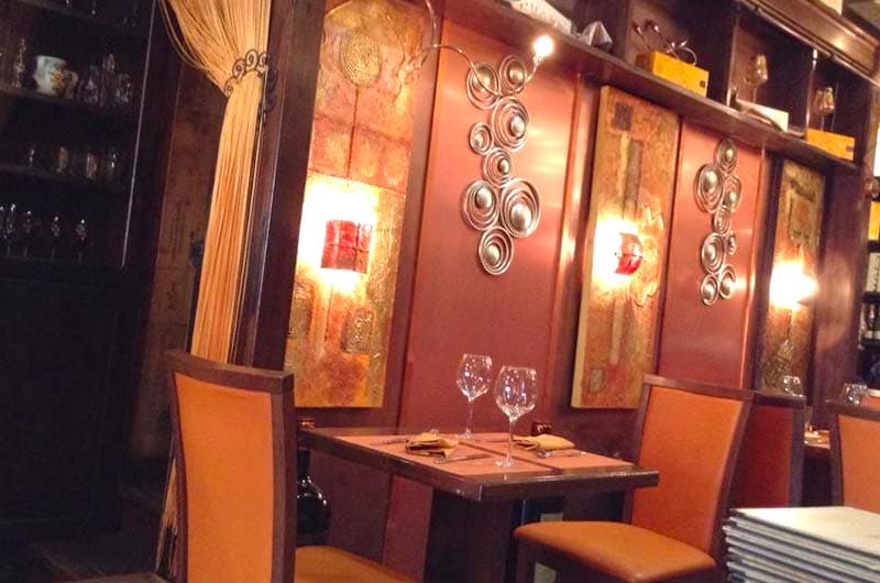 Tavolo apparecchiato con calici da vino tra sedie in pelle color ruggine e pannelli decorativi a spirali metalliche.