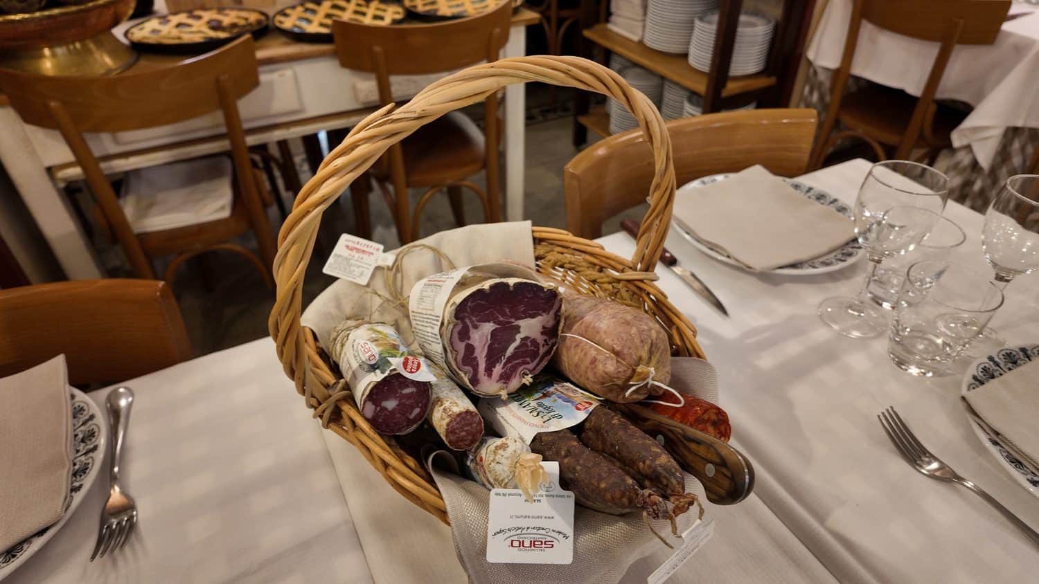 Cesto di vimini con salumi assortiti su un tavolo apparecchiato con tovaglia bianca.