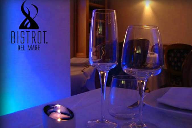 Calice da vino e flute su tavolo apparecchiato, con candela accesa e luce blu sullo sfondo, logo Bistrot Del Mare a sinistra.