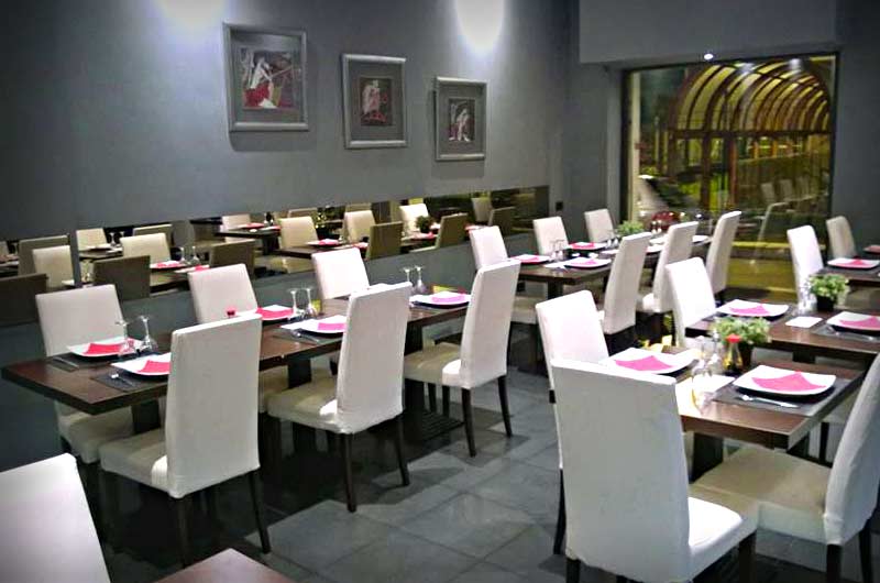 Sala con tavoli apparecchiati e tovaglioli rosa, sedie bianche alte e specchio lungo sulla parete grigia.