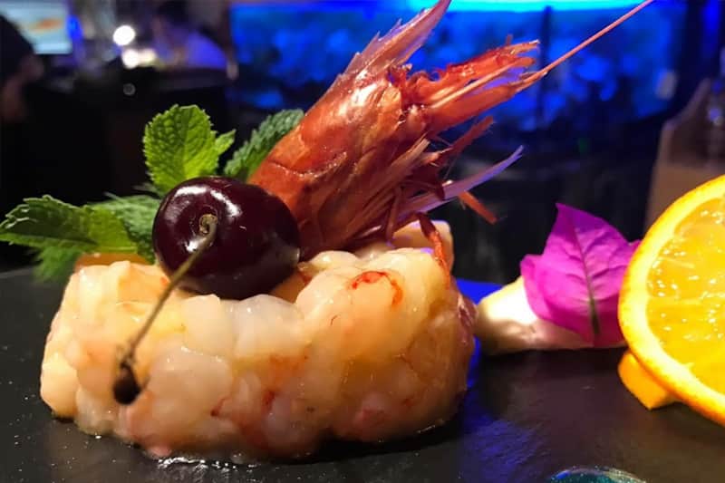 Tartare di gamberi su piatto scuro, sormontata da un gambero intero, una ciliegia e foglie di menta.