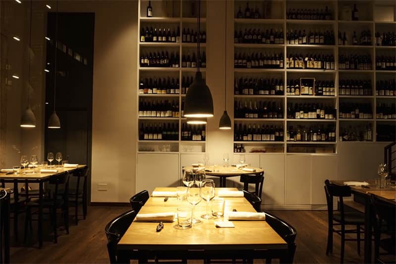 Sala da pranzo con tavoli in legno apparecchiati, lampade a sospensione e una parete di scaffali pieni di bottiglie di vino.