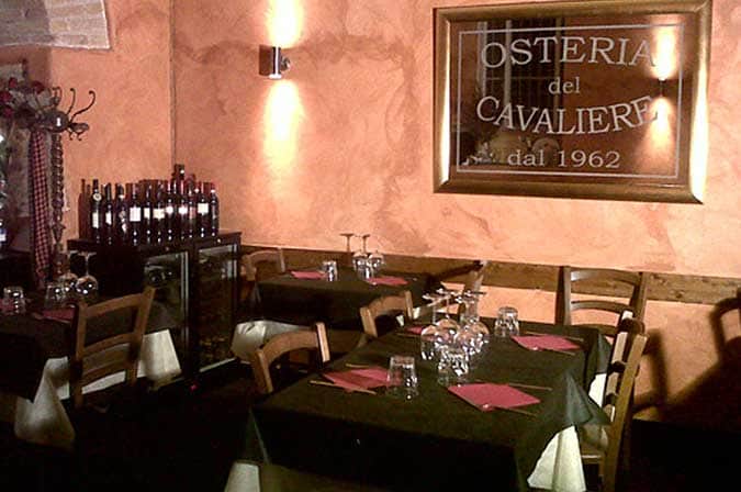 Sala di ristorante con tavoli apparecchiati, tovaglie scure e specchio inciso con la scritta Osteria del Cavaliere dal 1962.
