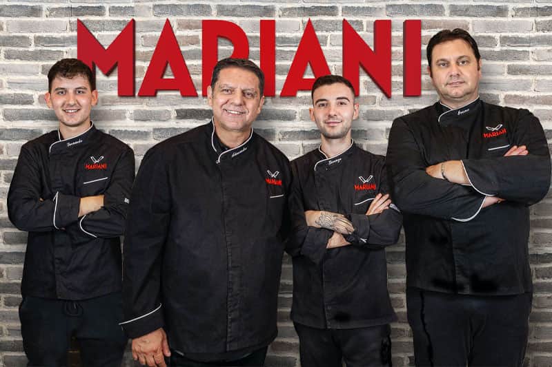 Quattro uomini in giacca da chef nera, braccia conserte, davanti a un muro di mattoni con la scritta rossa MARIANI.