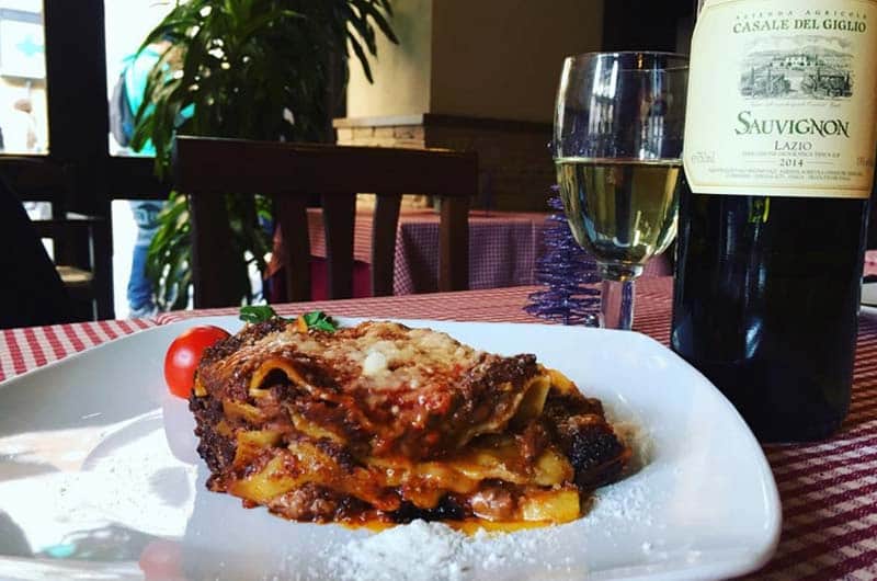 Porzione di lasagna al forno su piatto bianco, accanto a un bicchiere e una bottiglia di vino bianco.
