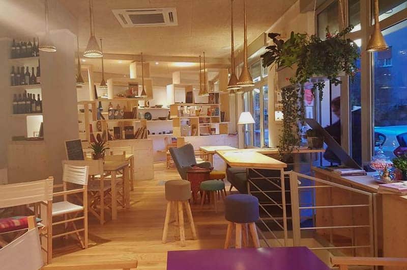 Sala con scaffali di libri e bottiglie, tavoli in legno, sgabelli colorati e lampade a sospensione dorate.