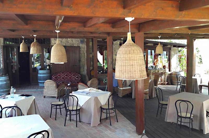Sala all'aperto sotto un pergolato in legno, con tavoli apparecchiati e lampade in vimini pendenti dal soffitto.