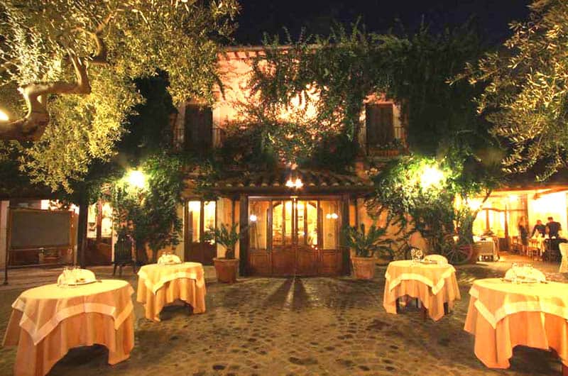 Tavoli con tovaglie arancioni apparecchiati all'aperto davanti a un edificio illuminato coperto di rampicanti, di sera.