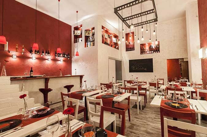 Sala di ristorante con bancone bar su parete rossa da un lato e tavoli con tovaglie rosse e sedie bianche dall'altro.