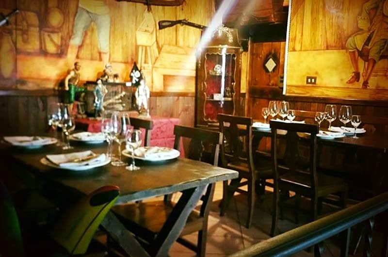Sala con tavoli apparecchiati di cristalli e tovaglioli, pareti dipinte con scene a tema marinaresco e vetrinetta in legno scuro.