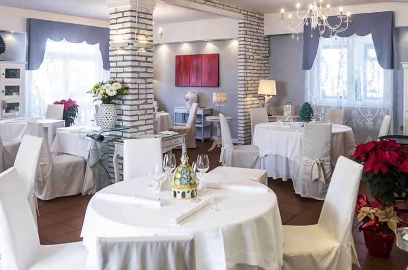 Sala con tavoli rotondi apparecchiati in bianco, colonne in mattoni e tende blu alle finestre.