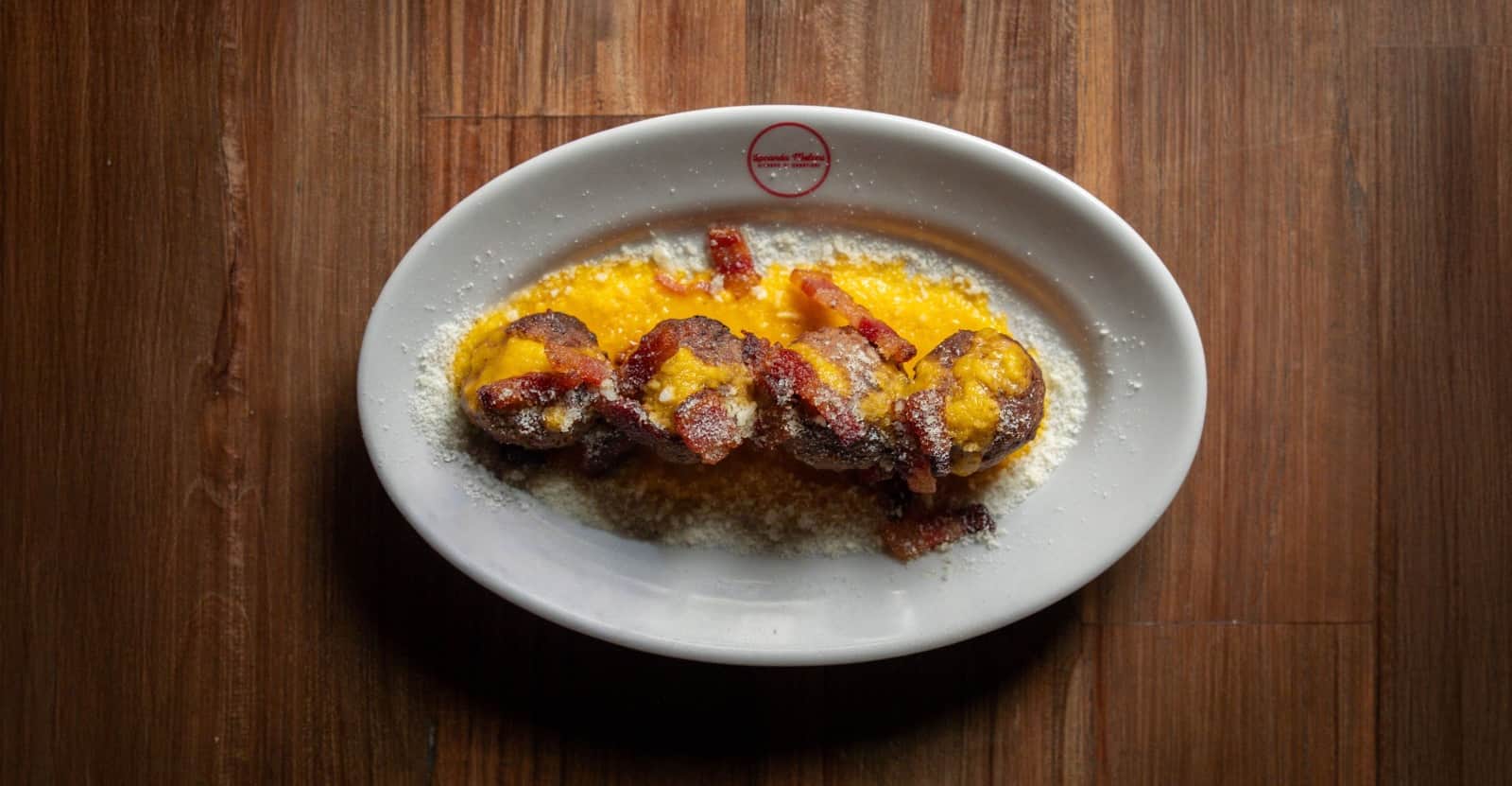 Piatto ovale con crema gialla, pezzi di carne scura e strisce di pancetta croccante, cosparso di formaggio grattugiato.