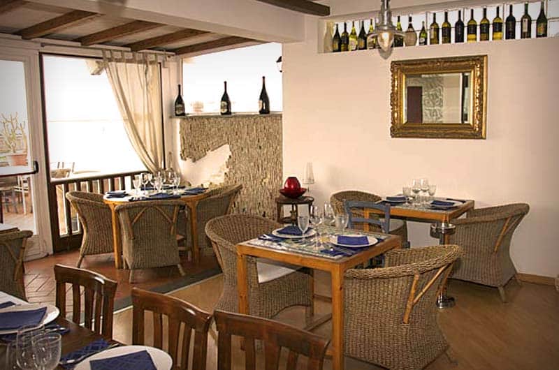 Sala con tavoli in legno e sedie di vimini, tovaglioli blu, bottiglie di vino su una mensola e specchio con cornice dorata.